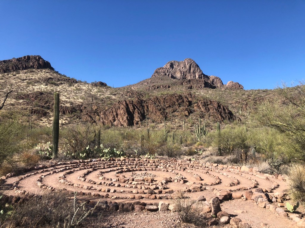 A stone labyrinth surrounded by desert vegetation with mountains in the background under a clear blue sky.