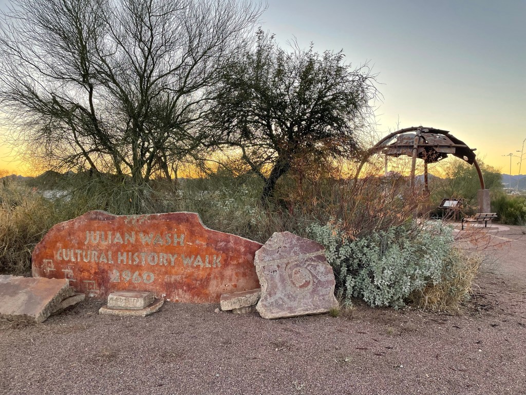 A sign marking the Julian Wash Cultural History Walk, with desert vegetation and a gazebo structure in the background during sunset.