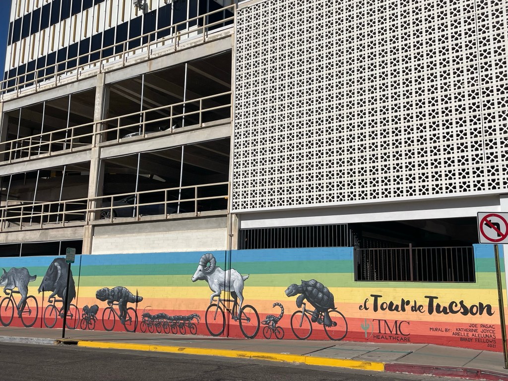 A colorful mural featuring various animals, including a ram, javelina, rabbit, tortoise, and dog, riding bicycles against a gradient background, with the text 'el Tour de Tucson' displayed prominently.