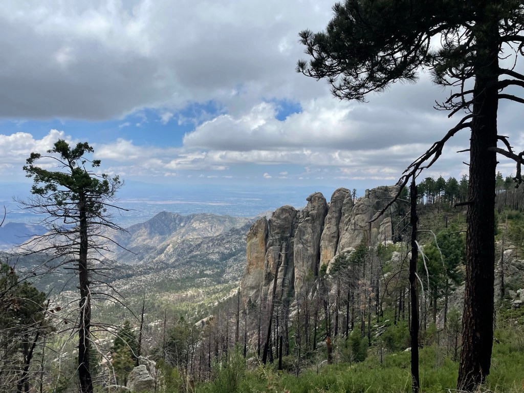 A scenic view of a mountainous landscape with rocky cliffs in the foreground and a valley extending into the distance, surrounded by trees and a partly cloudy sky.