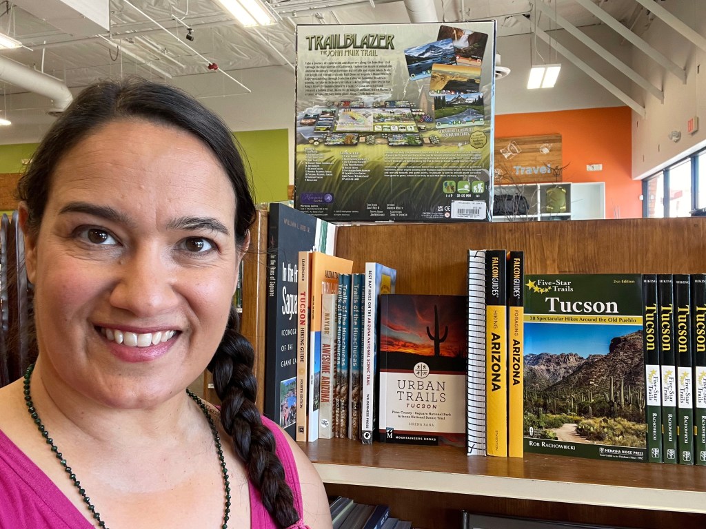 A person smiling in front of a shelf displaying several guidebooks, including 'Urban Trails: Tucson', in a bookstore setting.
